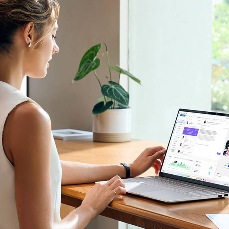 a young woman sitting at a desk near a window with her ASUS Vivobook S 15 laptop while using Copilot AI tool